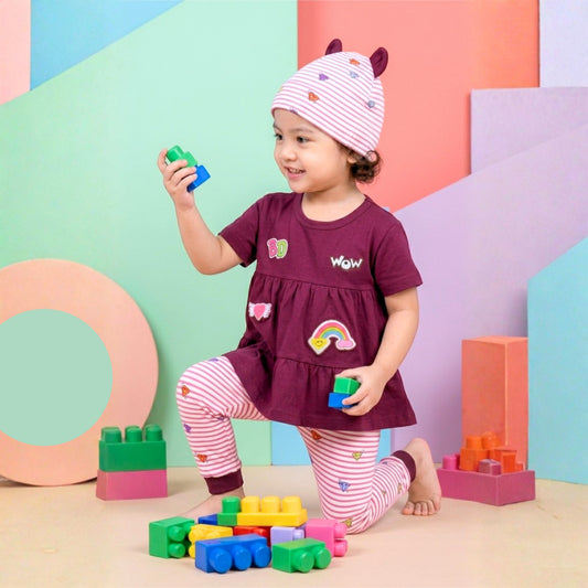 A toddler kneeling on a pastel geometric set, wearing a Babydrip 3 piece girl set that features a magenta short-sleeve Tunic Blouse with rainbow and heart graphic design details, and pink-and-white striped leggings, and a matching beanie hat, while playing with colorful building blocks.