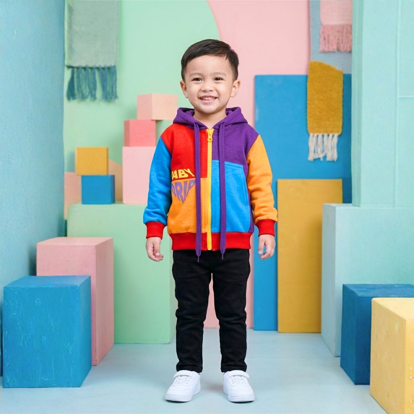 A young boy stands smiling in a colorful studio setting, wearing a Babydrip brightly color-block hoodie with purple, blue, orange, and red panels, with red ribbed cuffs and hem with a bright yellow zipper in the front of the sweatshirt. The boy has on a black pants, and white sneakers. The background features pastel colored geometric blocks with yellow, green and pink hanging textured textiles with frills at the end.