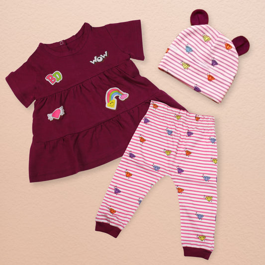 A baby girl's three-piece Babydrip clothing set laid flat on a light beige background. The set includes a Magenta short-sleeve ruffled tunic blouse with colorful graphic design appliques, matching pink and white striped pants with magenta ribbed cuffs with small multicolor diamond prints, all over the body of the pants and a pink and white striped beanie with magenta colored bunny ears.
