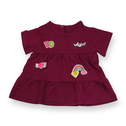 A Babydrip magenta short-sleeve tunic blouse with ruffled tiers, featuring embroidered patches including 'BabyDrip', (BD) 'WOW', a pink heart with angel wings, and a rainbow on a white background. Laid out flat on a white background.