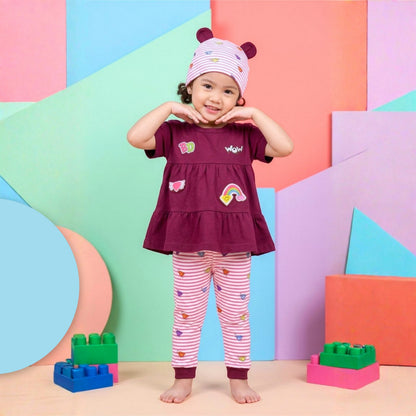 A little girl stands barefoot in a colorful studio setting, wearing a Babydrip short-sleeve Magenta colored tunic blouse with rainbow and heart graphic Design appliques, with matching pink-and-white striped leggings, and a magenta colored featured bunny ears with pink and white stripe beanie hat with small colorful diamond graphic details. The background consists of pastel geometric panels in pink, blue, green, and purple, with Lego toy blocks on the floor.