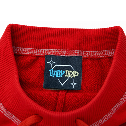 Close-up of a red ribbed knit terry cotton fabric waist with a navy blue sewn-in label reading BABY DRIP, in light blue and yellow text inside a white stitched with a drawstring nook at the front of the waist.