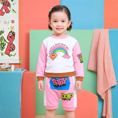 A young girl stands in a colorful studio setting wearing a Babydrip pink and white two-piece sweatsuit with cartoon graphics, including a rainbow on the front center of the chest of the long sleeve sweatshirt and the text 'BABY" on a blue kangaroo pocket at the front of the shorts and the word "DRIP" a little further down from the pocket.