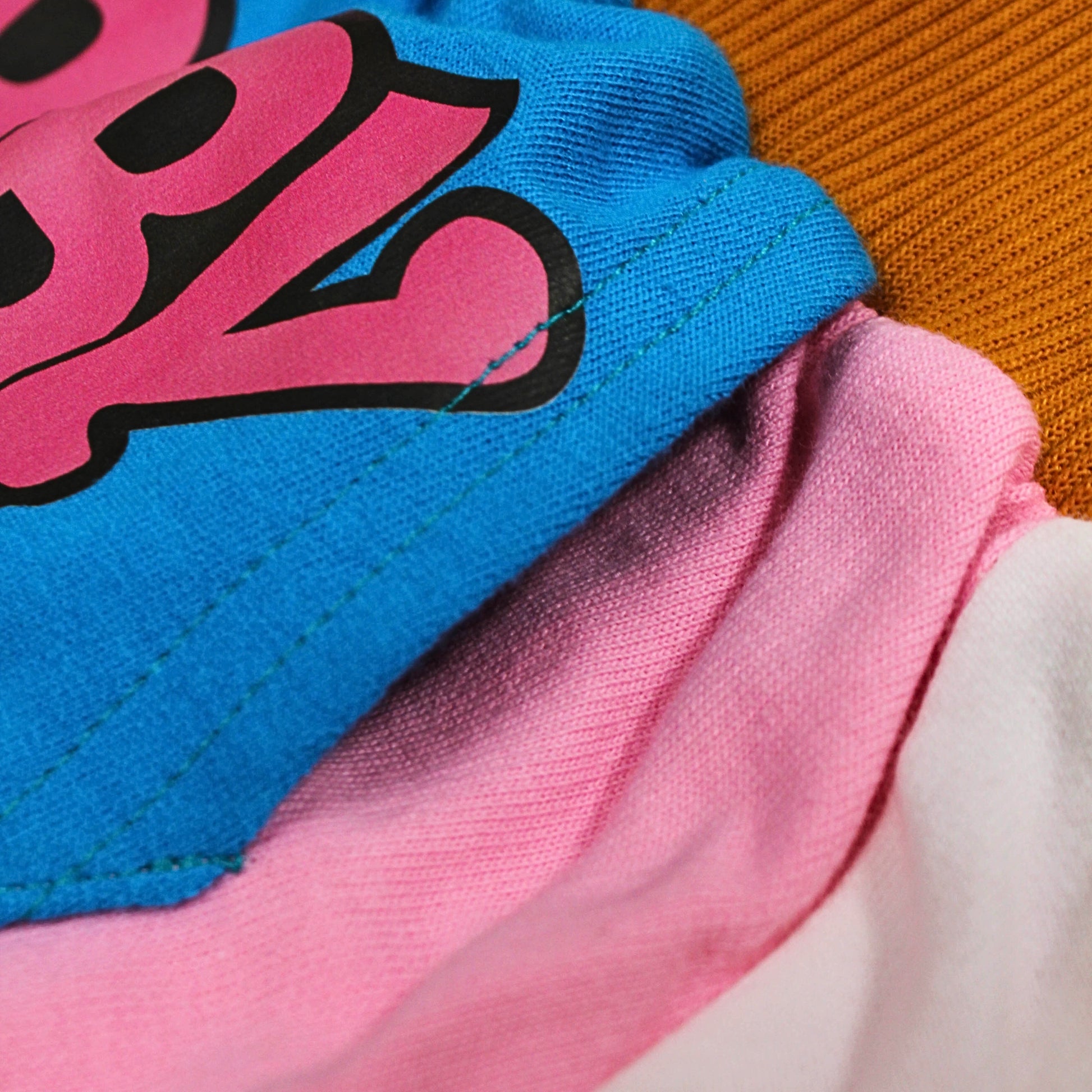 A close-up image of a Babydrip garment featuring a blue Kangaroo Pocket on a terry cotton garment showing a a part of the bright blue section with a large pink graphic outlined in black, layered over a pink section and a white section, with the brown ribbed elastic waistband fabric visible in the upper right.
