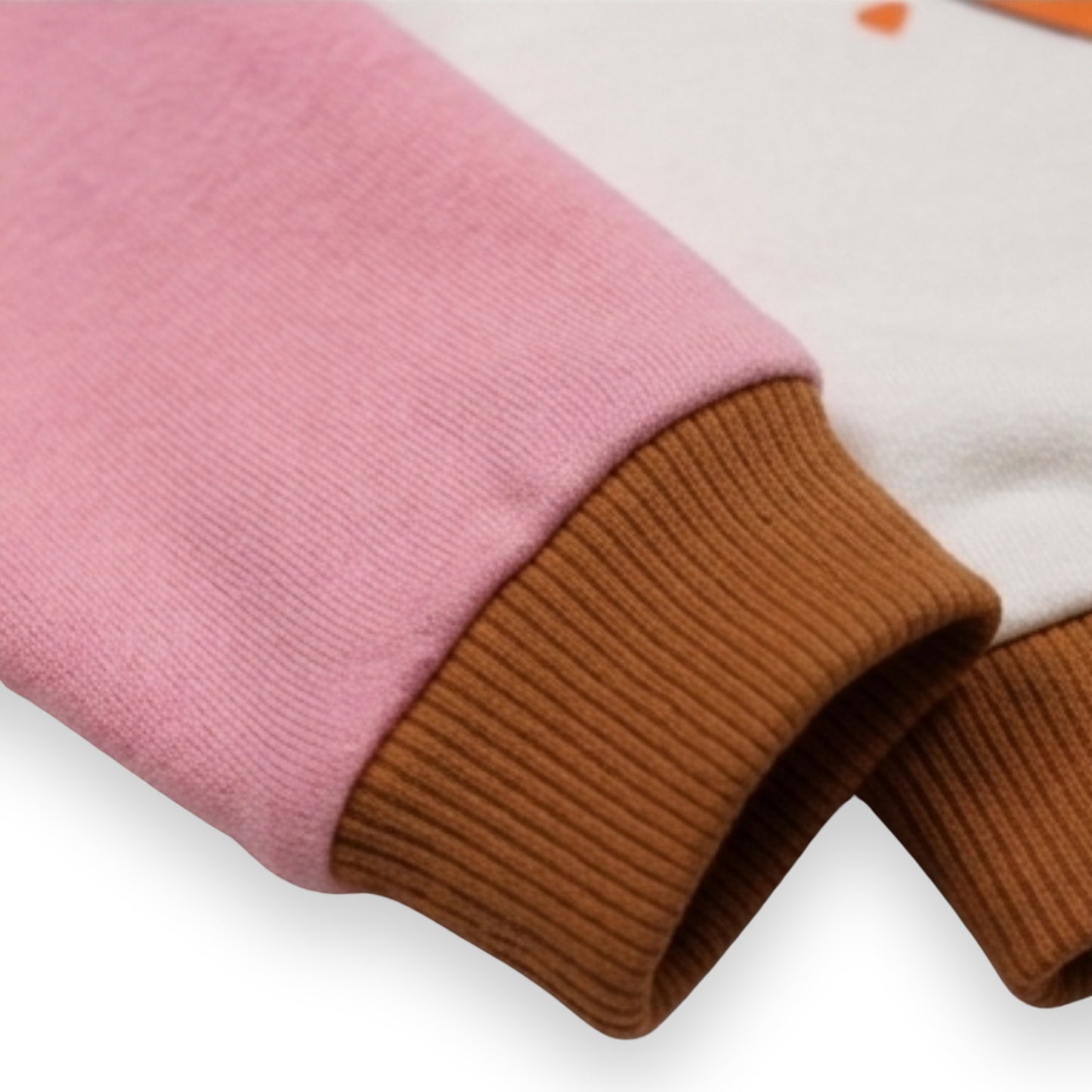 Close-up of a Babydrip terry cotton ribbed garment showing a pink sleeve with a brown ribbed cuff, a white body section, and part of a small orange graphic print laid out on a white background.