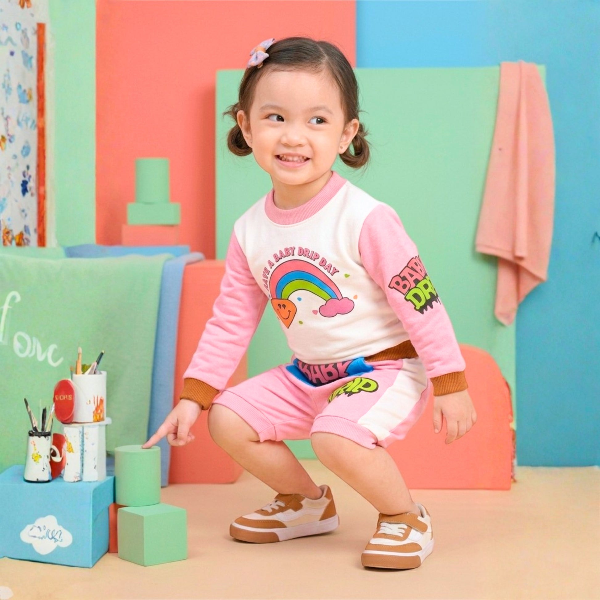 A young girl in a Babydrip pink and white two-piece terry cotton sweatshirt with rainbow, diamond, and cloud graphic print on the front of the shirt and pink and lime green graphic print on the right sleeve. The shorts is also pink with white stripe to the right side with cartoon graphics, "BABY' in pink text on a kangaroo blue pocket with the text 'DRIP" a little further from the front pocket. The little girl is posing and smiling in a studio decorated backdrop with pastel-colored building blocks.