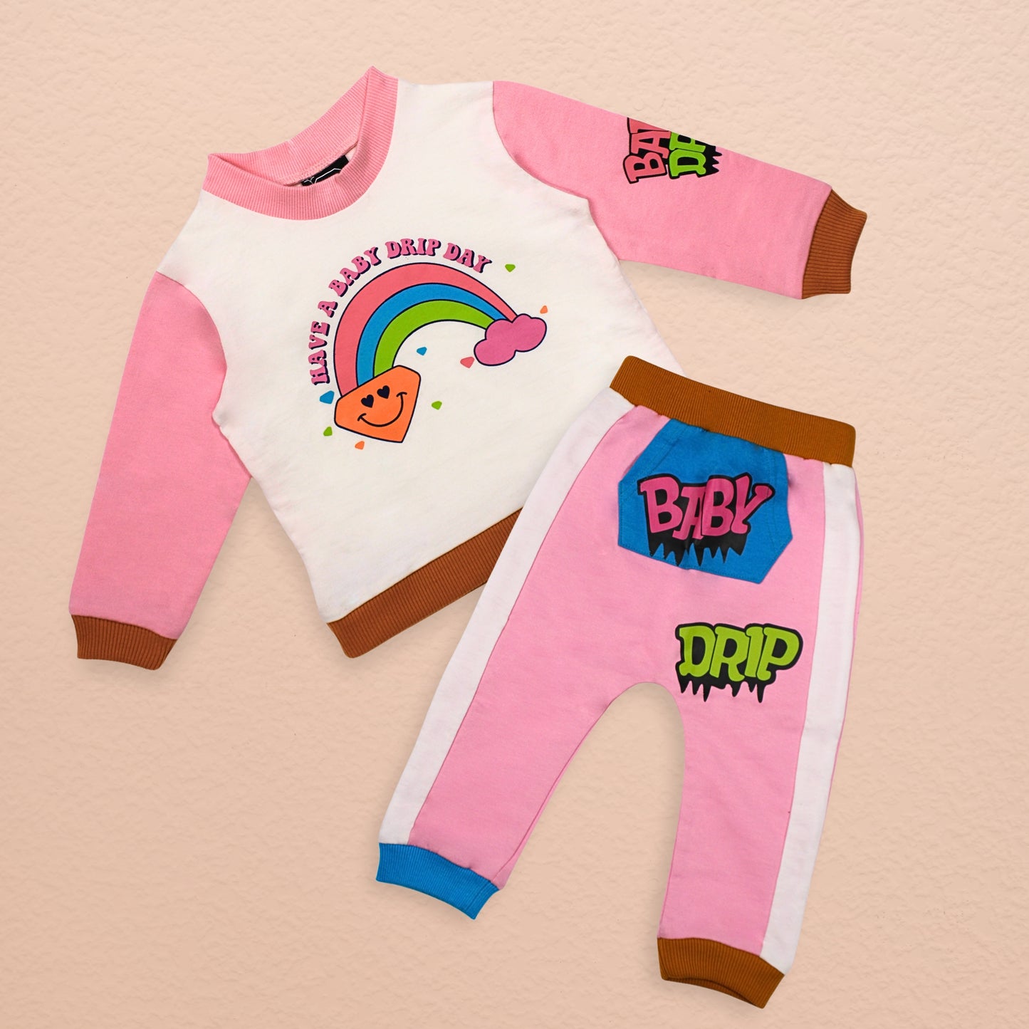 A Babydrip little girl two-piece sweatshirt outfit featuring pink long-sleeves and brown ribbed cuffs top with matching pants in pink and white, with brown cuffs and hem and white panels on each sides. The top has a colorful rainbow graphic and the text 'HAVE A BABY DRIP DAY', while the pants feature 'BABY DRIP' graphics with a blue Kangaroo pocket in the front, brown ribbed elastic waistband and blue and brown ribbed cuffs, laid out flat on a biege background.