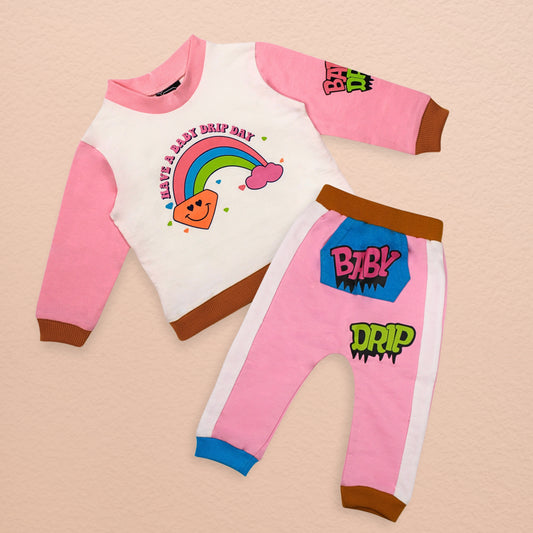 A Babydrip little girl two-piece sweatshirt outfit featuring pink long-sleeves and brown ribbed cuffs top with matching pants in pink and white, with brown cuffs and hem and white panels on each sides. The top has a colorful rainbow graphic and the text 'HAVE A BABY DRIP DAY', while the pants feature 'BABY DRIP' graphics with a blue Kangaroo pocket in the front, brown ribbed elastic waistband and blue and brown ribbed cuffs, laid out flat on a biege background.