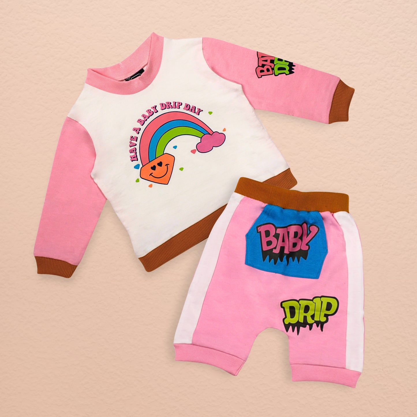 A Babydrip two-piece sweatshirt and shorts set outfit featuring pink long-sleeves and neck with a white body with a colorful rainbow and text "HAVE A BABY DRIP DAY" with brown hems and matching shorts that features a blue kangaroo front pocket with the text "BABY" at the front of the shorts with the text "DRIP" a little further down on the front of the shorts, also featuring a elastic waist band for easy pull up and down access laid out on cream colored background.