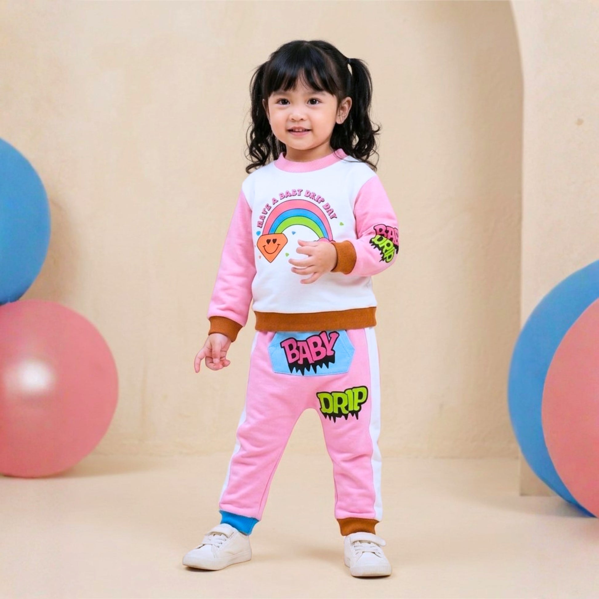 A young Asian girl wearing a Babydrip pink and white 2 piece terry cotton sweatshirt with rainbow, diamond, cloud graphic print on the front and pink and lime green graphic print on the right sleeve. The pants is also pink with white stripes to the sides with cartoon graphics, "BABY' in pink text on a kangaroo blue pocket with the text 'DRIP" further down from the front pocket, wearing a white sneakers. Posing and smiling in a studio decorated with pastel-colored balls.