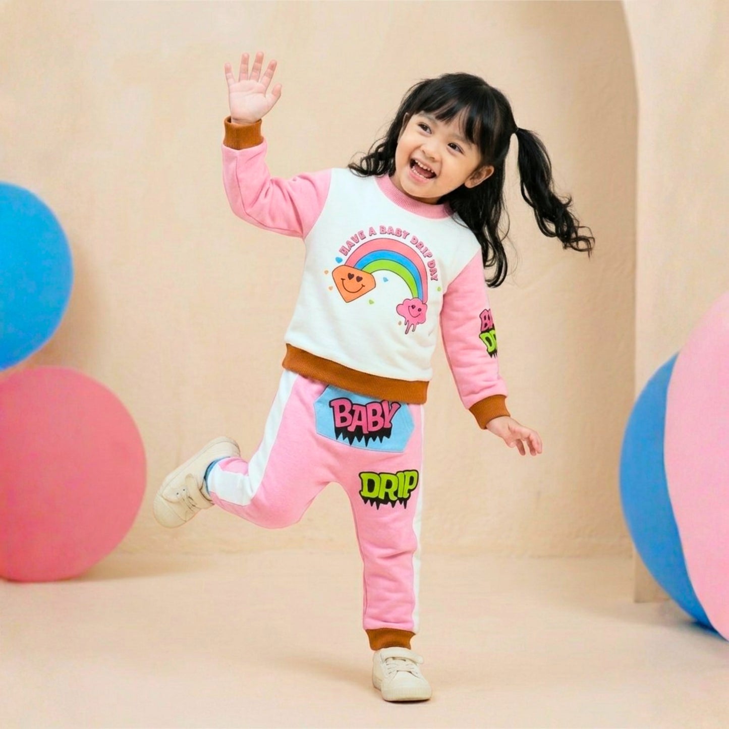 A little girl wearing a Babydrip pink and white two-piece terry cotton sweatshirt with rainbow, diamond, and cloud graphic print on the front and pink and lime green graphic print on the right sleeve. The pants is also pink with white stripes to each side with cartoon graphics, "BABY' in pink text with black outline on a kangaroo blue pocket with the text 'DRIP" a little further from the front pocket of the pants. Wearing a white sneakers, posing playfully in a studio decorated with pastel-colored balls.