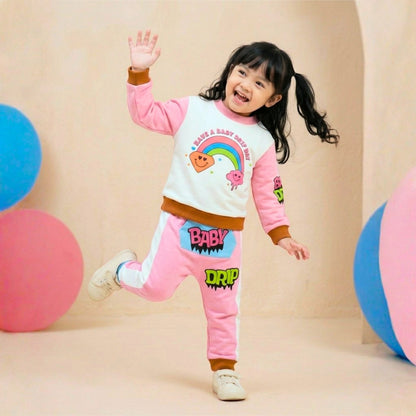 A little girl wearing a Babydrip pink and white two-piece terry cotton sweatshirt with rainbow, diamond, and cloud graphic print on the front and pink and lime green graphic print on the right sleeve. The pants is also pink with white stripes to each side with cartoon graphics, "BABY' in pink text with black outline on a kangaroo blue pocket with the text 'DRIP" a little further from the front pocket of the pants. Wearing a white sneakers, posing playfully in a studio decorated with pastel-colored balls.