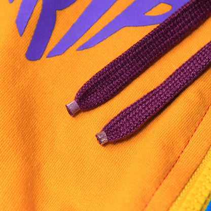 A Babydrip Close-up image of a bright orange garment with purple drawstrings with plastic tips and a part of a purple graphic print on the yellow section of the garment.