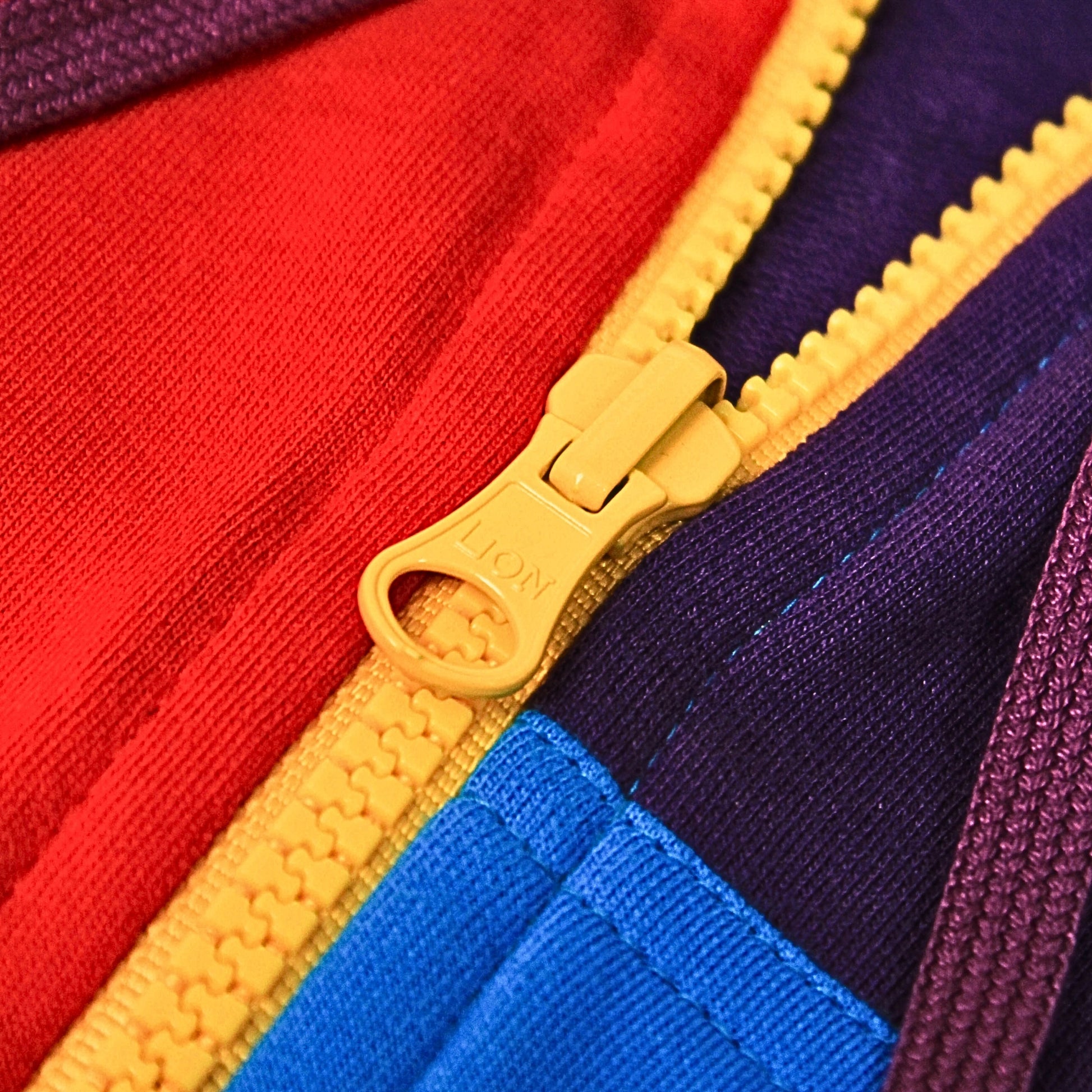 A Babydrip sweatshirt close-up image of the colorful zippered front section of the garment showing a yellow zipper pull with the brand name 'LION' embossed on it, alongside red, blue, and purple fabric panels.