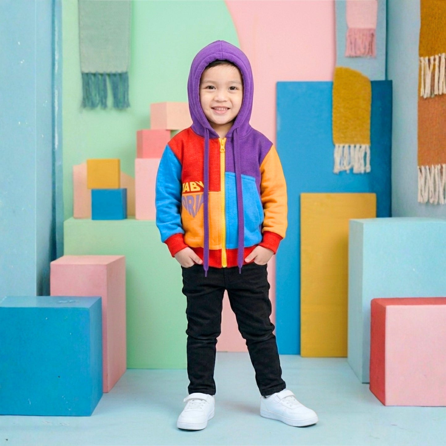 A young boy stands smiling with his hands in his pockets in a colorful studio setting backdrop, wearing a Babydrip sweatshirt hoodie that is brightly color-blocked with the hood over his head. The sweatshirt has purple, blue, orange, and red panels with a bright yellow zipper to the front. The boy is wearing a black pants, and white sneakers. The background features pastel geometric blocks and hanging textured textiles.